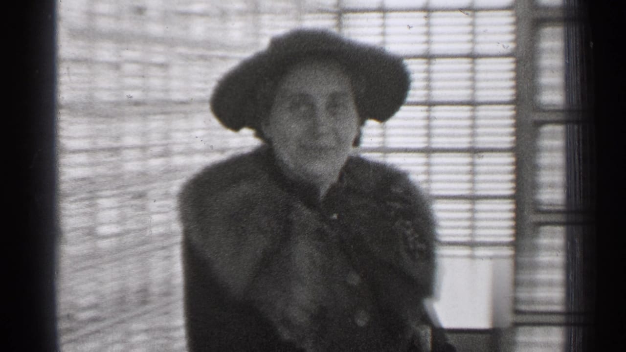 In 1944 San Francisco home-movie footage, an adult woman in a hat and fur stole stands by a window with blinds.