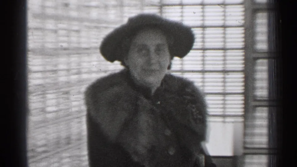 In 1944 San Francisco home-movie footage, an adult woman in a hat and fur stole stands by a window with blinds.