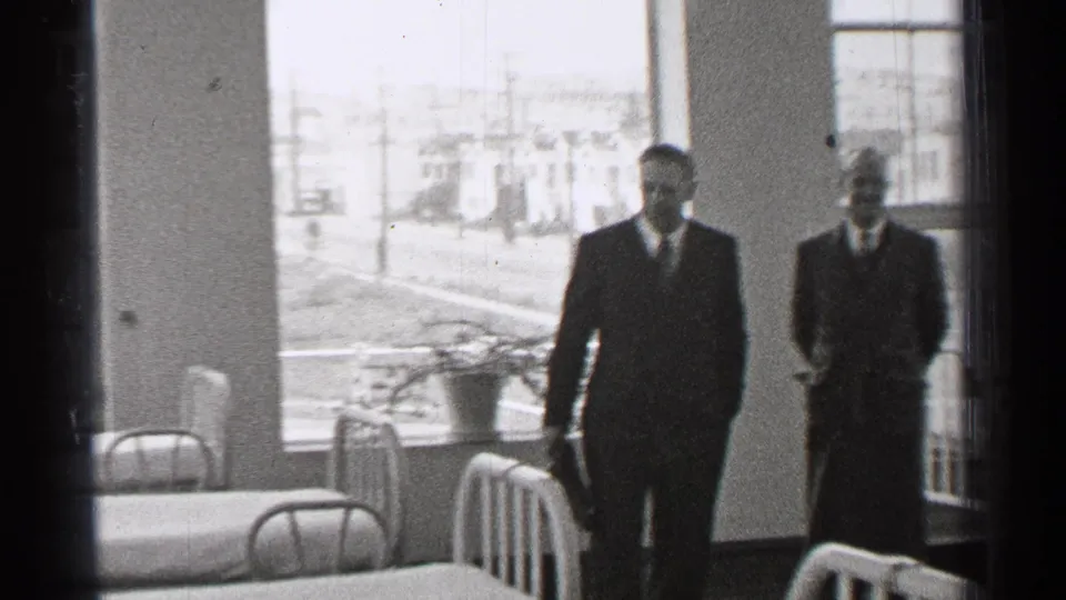 Two adult men in suits appear inside a 1944 San Francisco hospital ward with metal beds, white linens, potted plant, and street-facing windows.