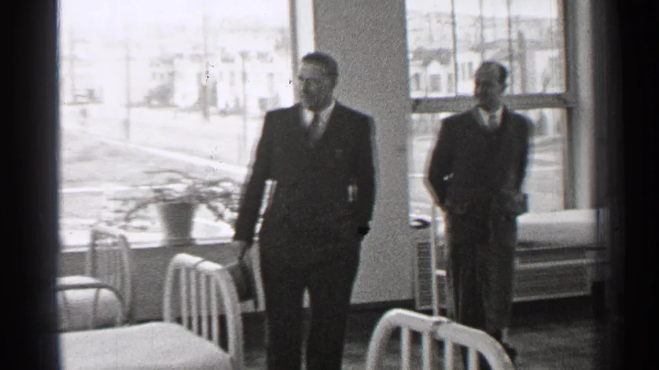 In the San Francisco ward, one suited man walks between metal beds while another stands by large windows overlooking a residential street.