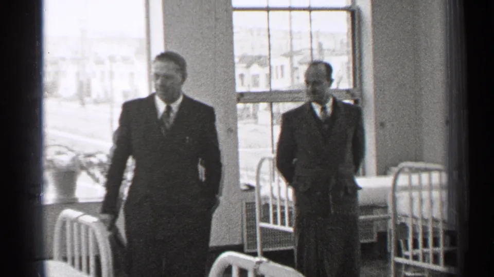 Two suited men are framed among metal hospital beds in the 1944 San Francisco ward, with bright windows and houses visible outside.