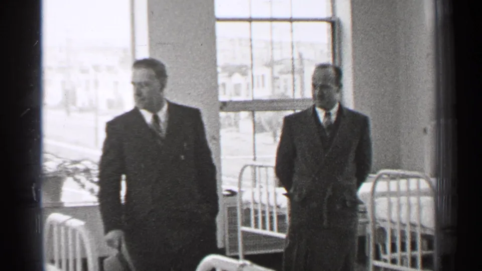 In 1944 San Francisco footage, two well-dressed men move through a hospital ward with metal beds and large windows behind them.