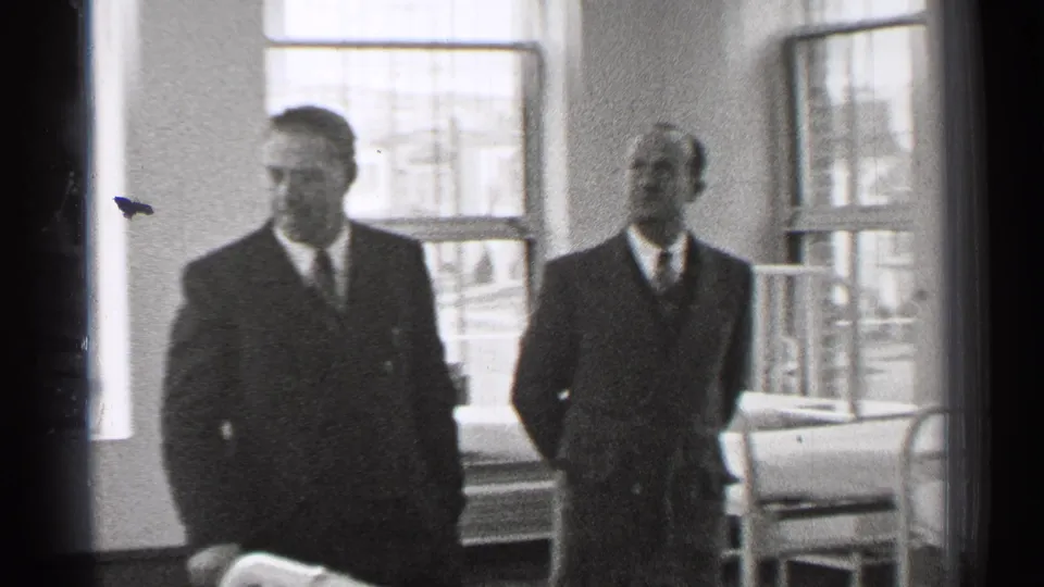The two suited men remain in the hospital ward, framed by tall windows and metal bed frames in black-and-white 8mm footage.