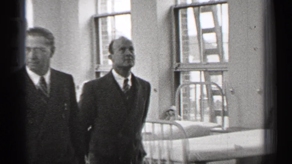 A closer 1944 San Francisco frame shows two suited men near a hospital bed, with a ladder visible by the tall windows.