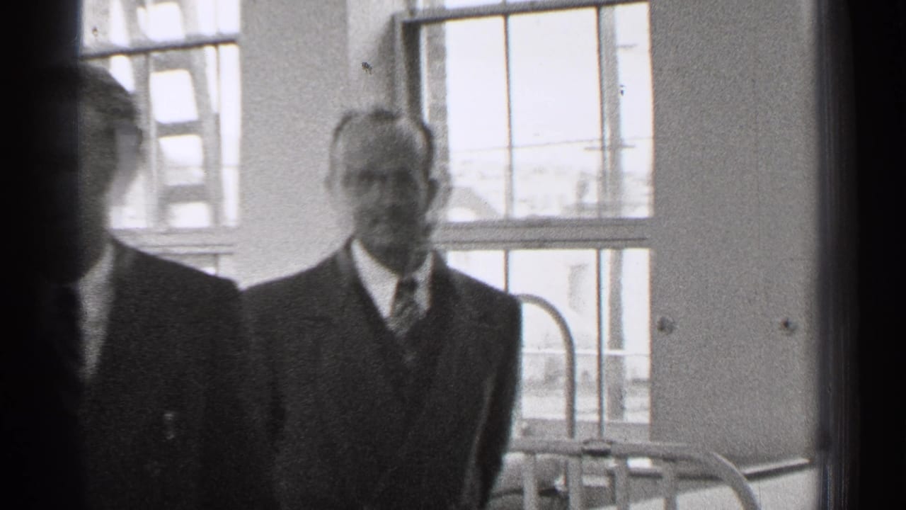 Two men in suits stand near large windows inside a room.