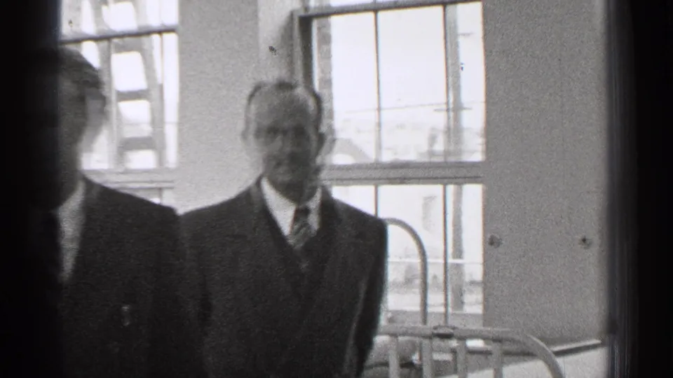 In the supported 1944 San Francisco home-movie context, two suited men stand in a bright interior near tall windows and plain walls.