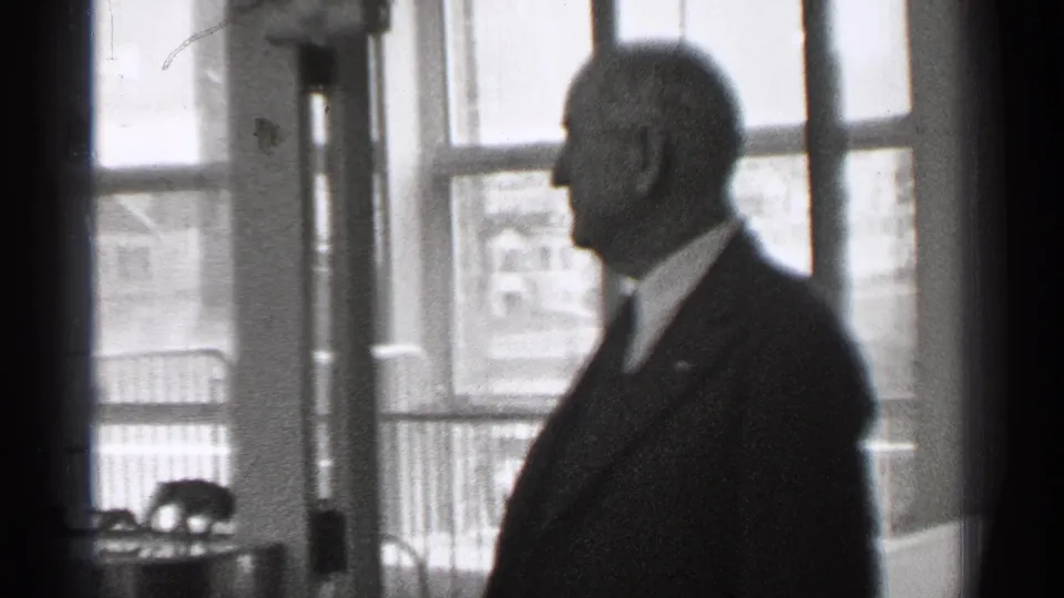 1944 San Francisco home-movie frame showing a man in a dark suit in profile beside tall windows in a private interior.
