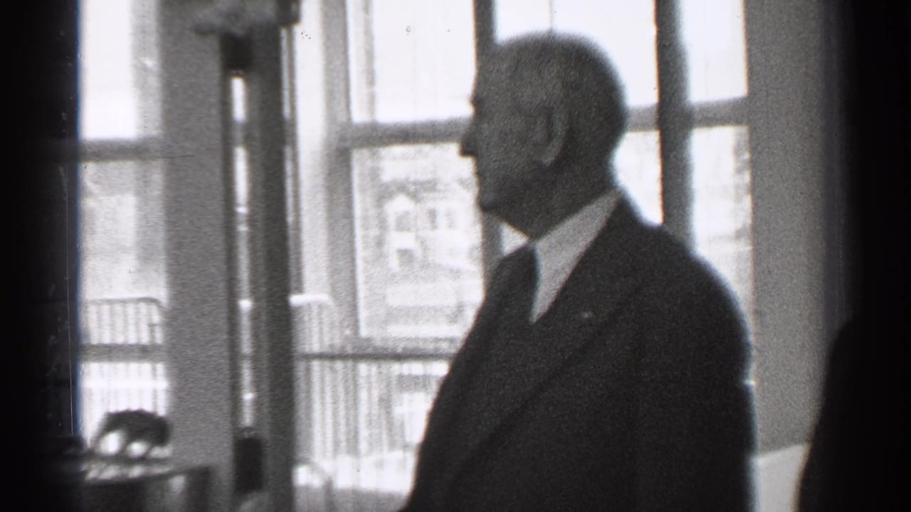 In 1944 San Francisco home-movie footage, a suited man stands in profile near tall windows inside a dim interior.