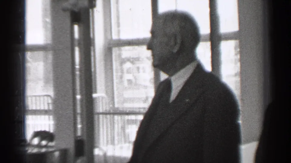 In 1944 San Francisco home-movie footage, a suited man stands in profile near tall windows inside a dim interior.