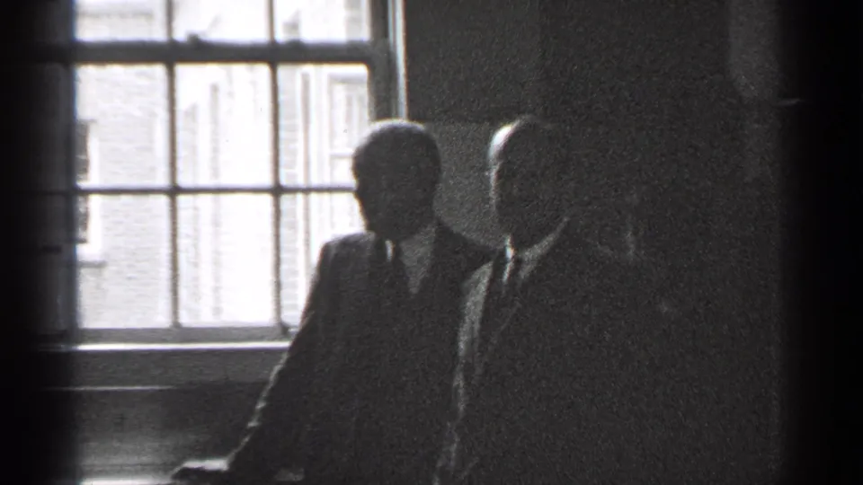 Men in suits and another adult appear in a dim San Francisco interior, backlit by a large window.