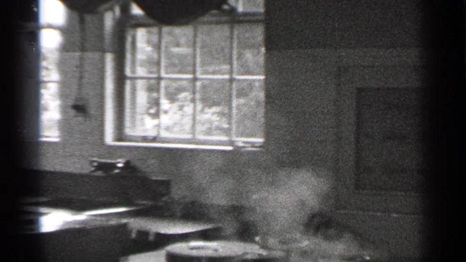 The camera looks across a kitchen or service counter toward paned windows, with a blurred person moving through the foreground.