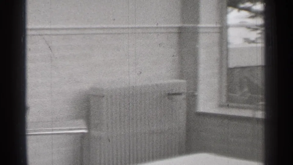 1944 San Francisco 8mm frame showing a radiator below a plain wall, with a small framed picture and window corner nearby.