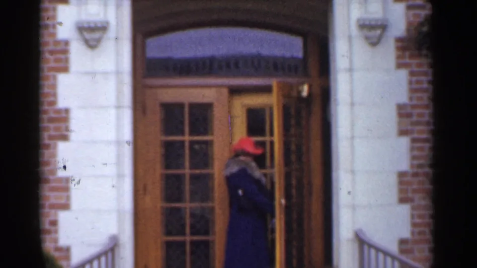 In the 1944 San Francisco clip, a woman in a red hat and dark coat is seen at a wooden double door set in brick and pale stone.