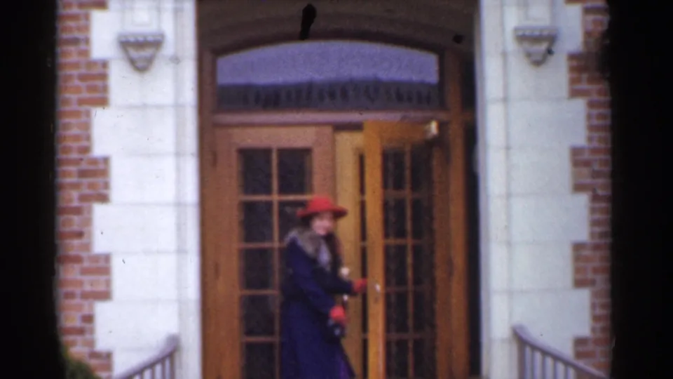 A woman in a red hat and dark coat moves through the wooden doorway of a brick and pale stone building in the 1944 San Francisco footage.