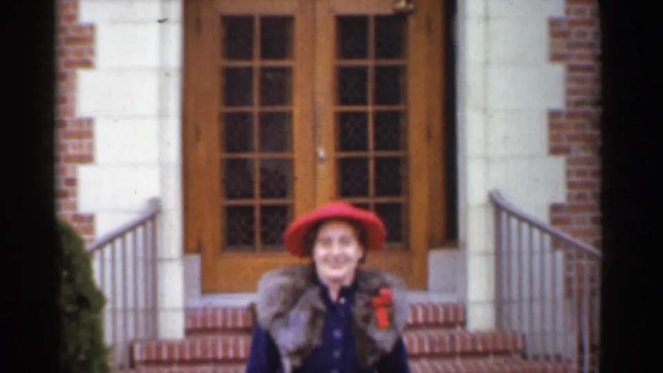 A smiling woman in a red hat and fur-collared coat nears the camera outside a San Francisco residence in 1944 home-movie footage.