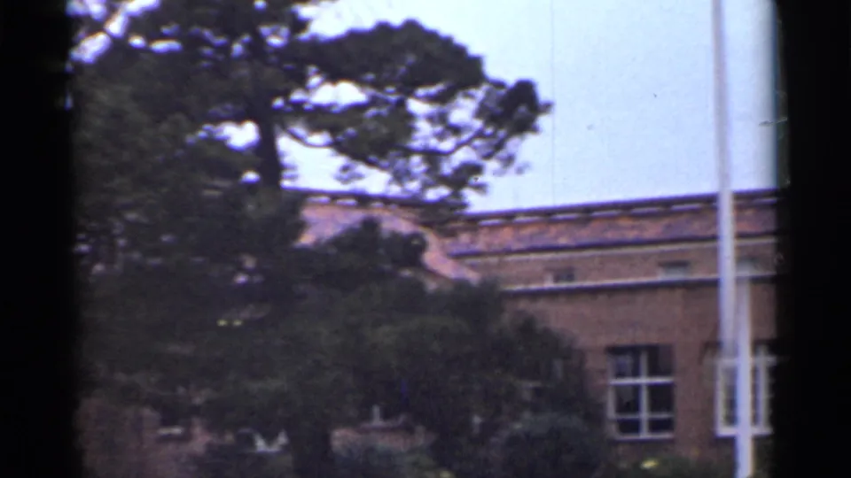 1944 San Francisco frame showing trees, shrubs, a brick building facade, windows, and a white pole on institutional grounds.