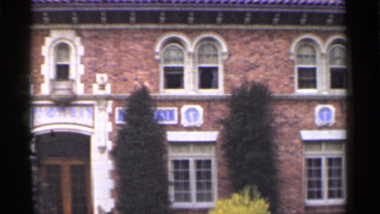 1944 San Francisco home-movie frame of a red brick facade with arched windows, pale stone entry trim, blue emblems, and trees.