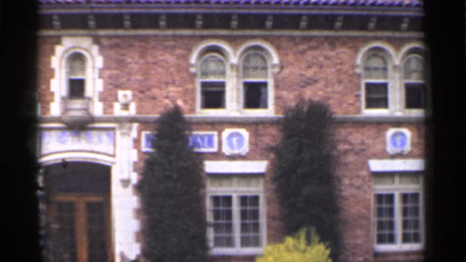 1944 San Francisco home-movie frame of a red brick facade with arched windows, pale stone entry trim, blue emblems, and trees.