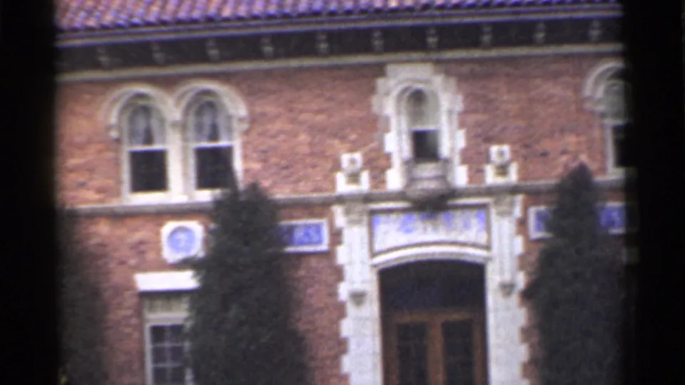 1944 San Francisco frame showing the building entrance area with arched windows, white trim, blue emblems, and evergreen trees.