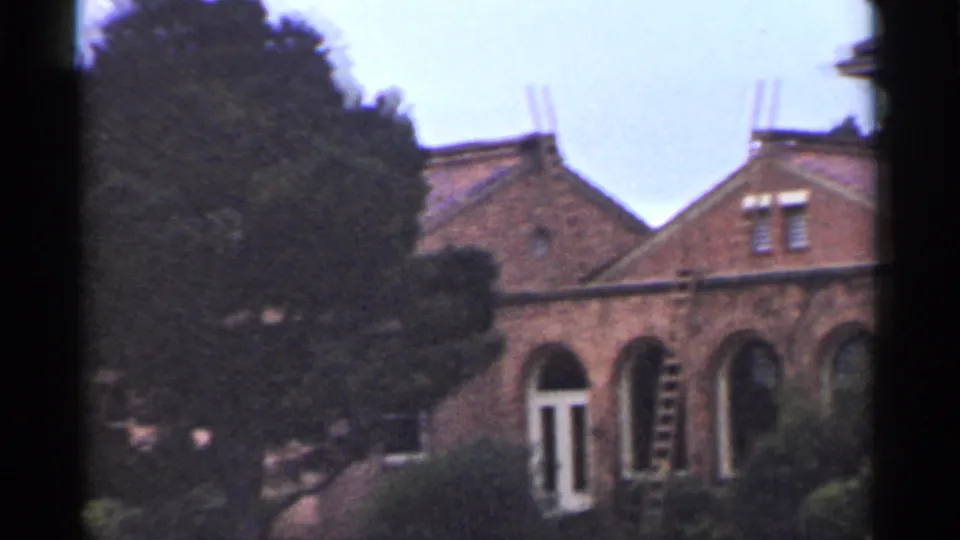 1944 San Francisco 8mm frame of a brick building wing with arched openings, tiled roof, large tree, and a ladder leaning near the wall.