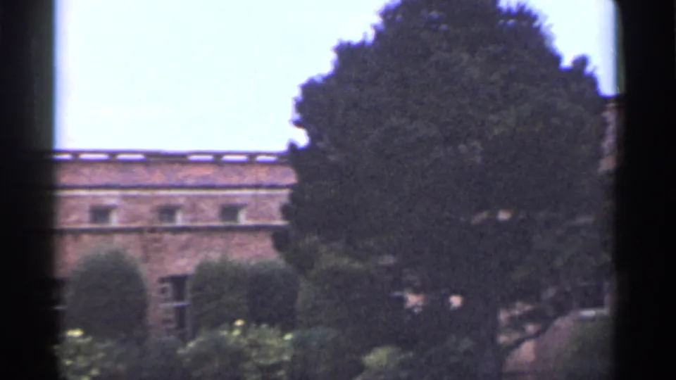 1944 San Francisco 8mm frame showing a long brick wall, small windows, clipped shrubs, garden plantings, and a large evergreen tree in front.