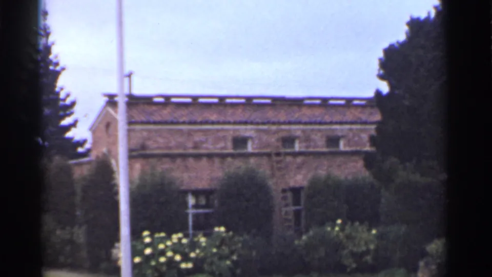 1944 San Francisco 8mm frame of a brick building side with small windows, tiled roof, shrubs, flowering plants, a flagpole, and trees.