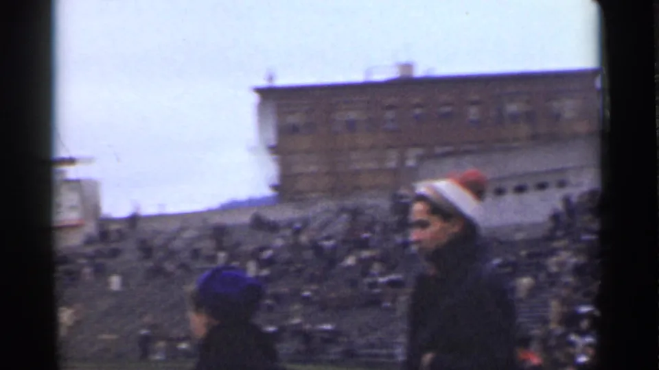 1944 San Francisco home-movie frame with foreground spectators in knit hats and crowded bleachers behind them.