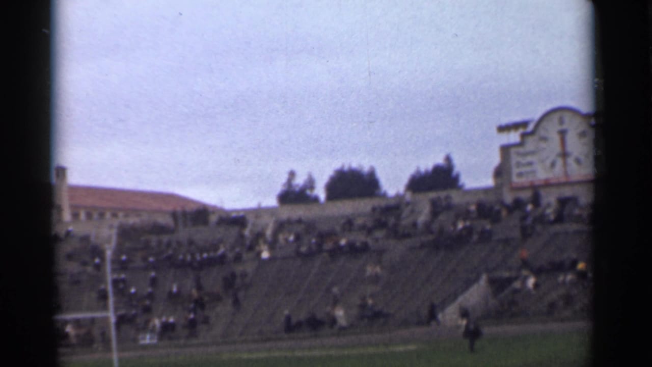 Crowded stadium stands with goalposts and a red-tiled building beyond the field.