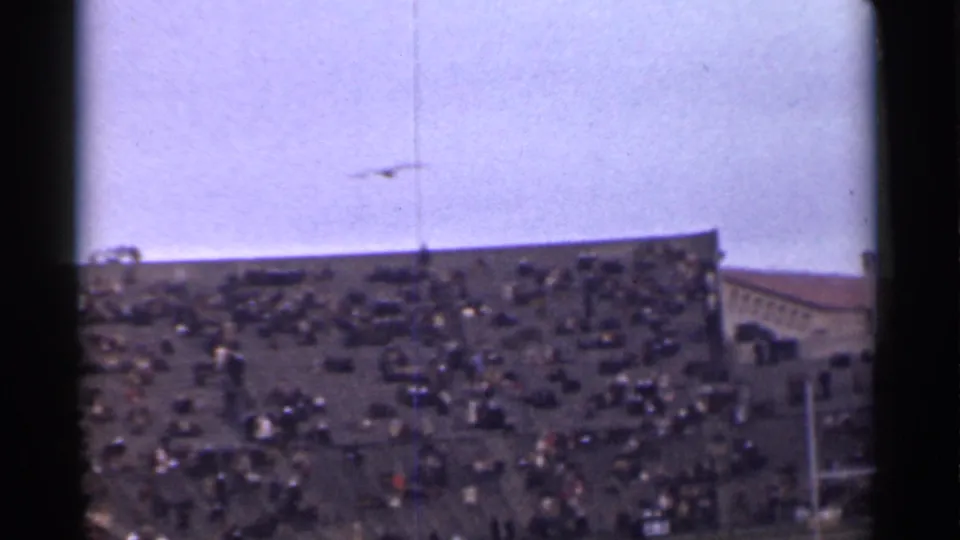 Faded 8mm frame of a crowded San Francisco stadium, with spectators spread across the stands and a goalpost near the field.