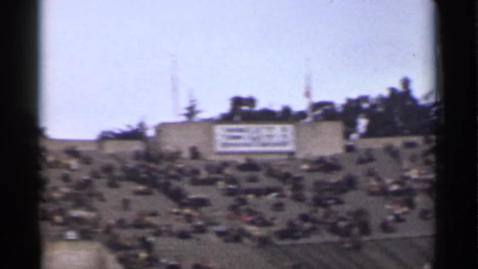 1944 San Francisco 8mm frame with crowded stadium seating, a light-colored field-side structure, flags or poles, and trees beyond.