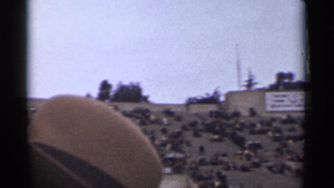 Large outdoor crowd seated on a hillside with distant structures and a dark foreground shape.