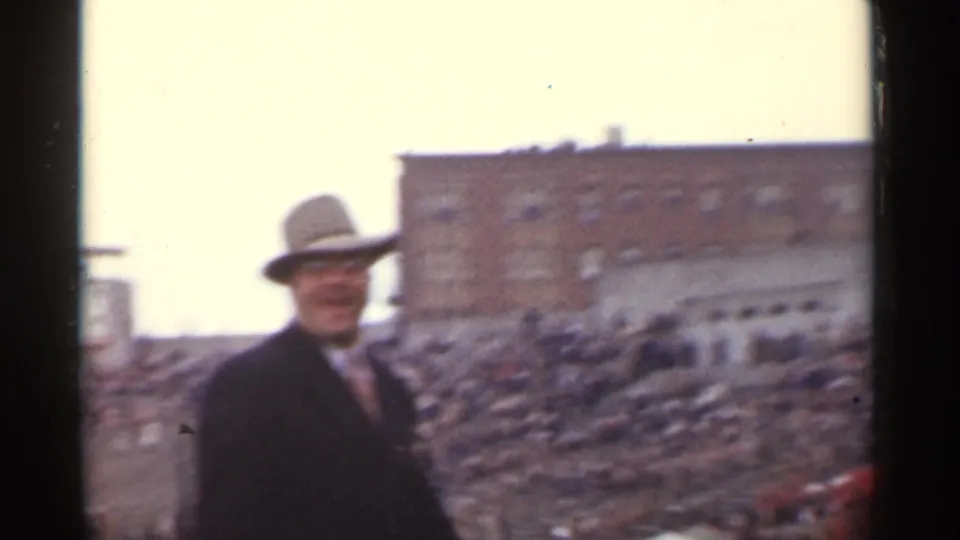 A suited man in a fedora appears in front of a stadium-like crowd in the San Francisco 1944 home-movie scene.