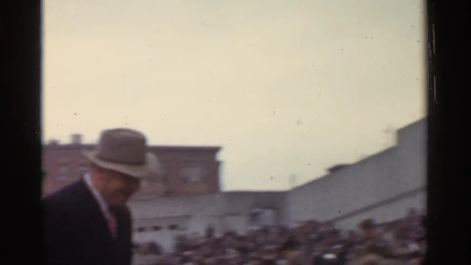 The fedora-wearing man is framed at the edge of a crowded outdoor event, with rows of spectators and nearby buildings behind him.