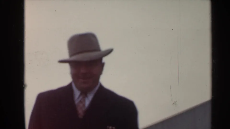 1944 San Francisco frame of a smiling man in a fedora, suit, and tie walking toward the camera with stadium structure behind him.