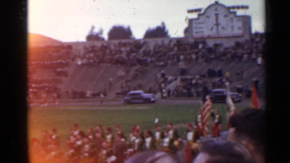 1944 San Francisco stadium view showing spectators in stands, flags at field level, parked cars, grass, and a large clock-tower-like structure.