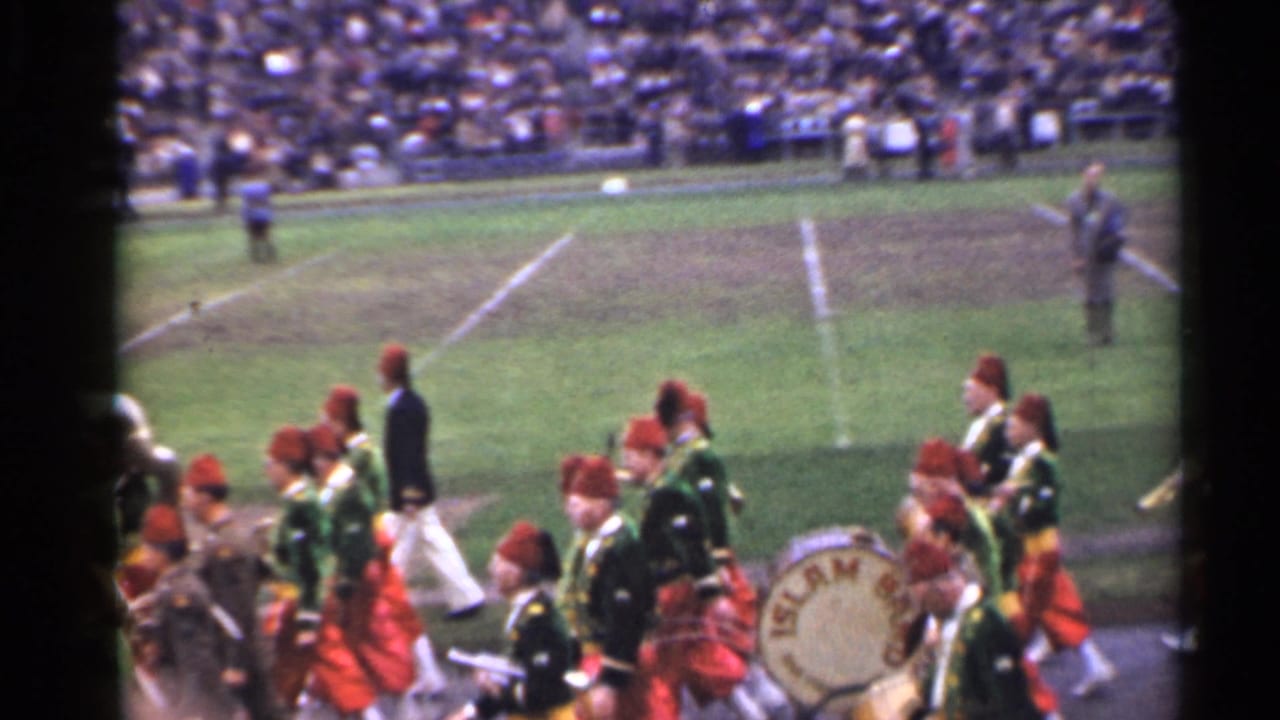 Marching band in red and green uniforms crossing a football field before crowded stands.