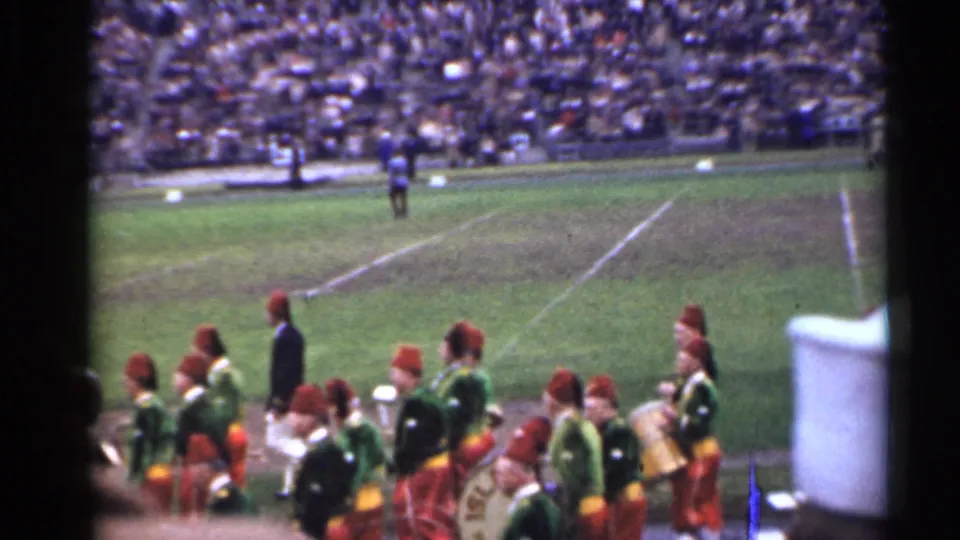 Spectator-shot 8mm frame of a 1940s San Francisco marching band moving across a football field with crowded stands behind.