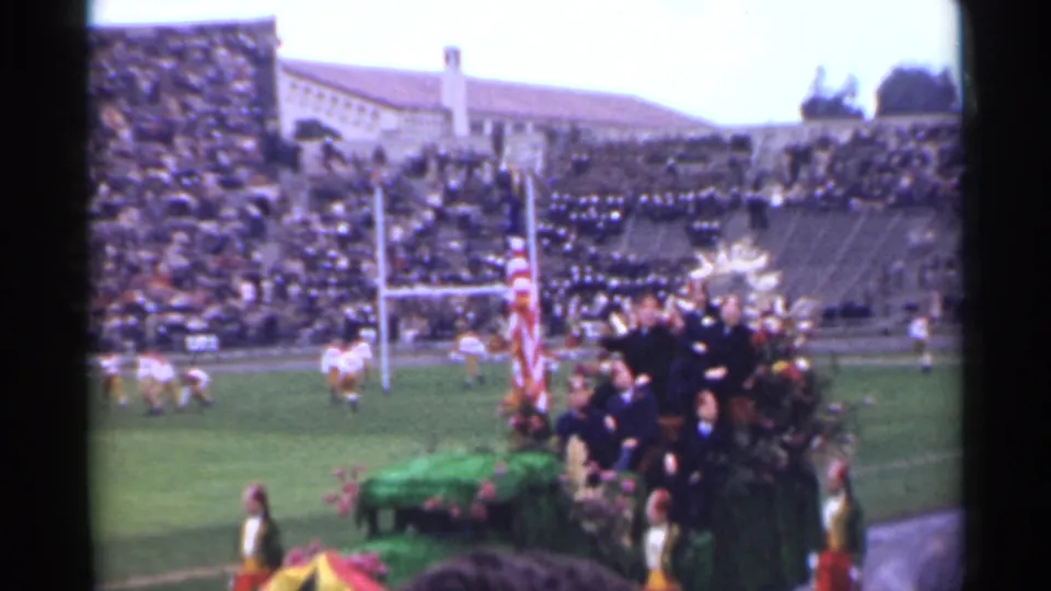 Color 8mm frame of a 1944 San Francisco football game with goalposts, players, spectators, and a decorated platform near the sideline.