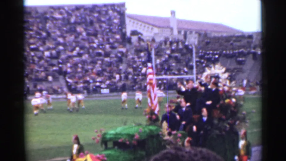 1944 San Francisco football stadium frame with players, crowd-filled stands, goalposts, an American flag, and a floral ceremonial platform.