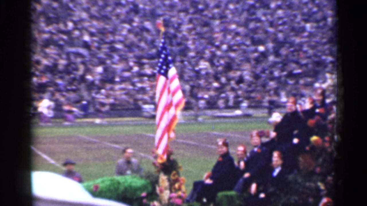 American flag near seated figures on a football field with crowded stadium stands behind.