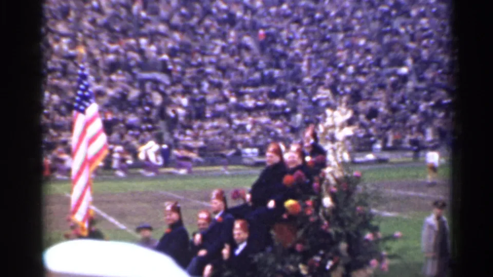 San Francisco 1944 frame of a field ceremony with an American flag, seated people, and dense football-stadium crowd behind them.
