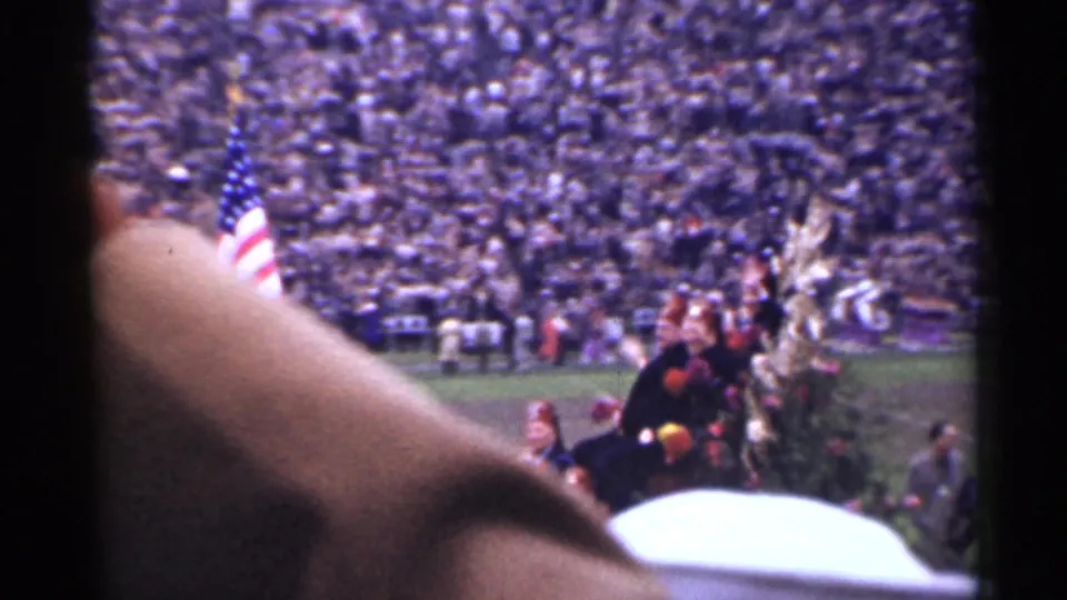 Obstructed 8mm home-movie view from the stands, showing the American flag, field guests, and crowded San Francisco football stadium.