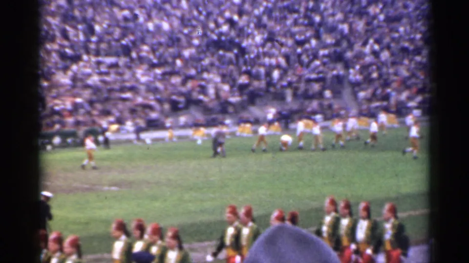 1944 San Francisco 8mm frame showing a high school football game from the stands, with band members in the foreground and a dense crowd beyond the field.