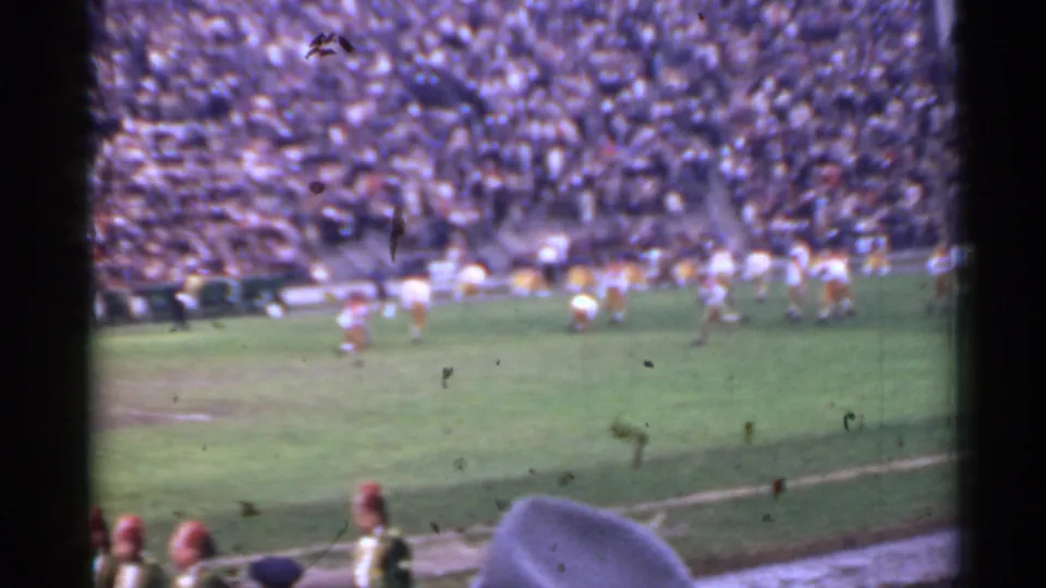 Home-movie 8mm view of a 1944 San Francisco football game, with soft field action, crowded bleachers, and a marching band member partly visible in front.