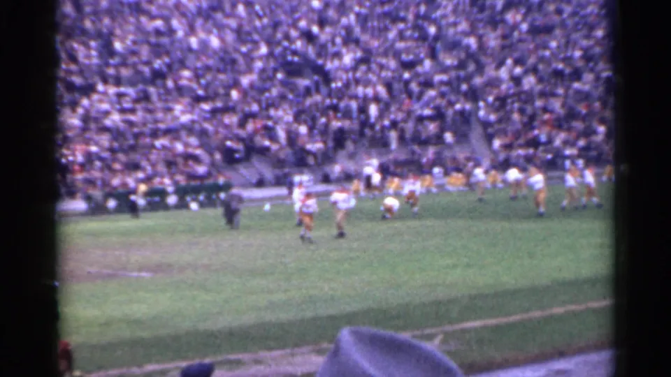 1944 San Francisco 8mm stadium view showing high school football players, a sideline official, packed spectators, and a nearby viewer partly blocking the frame.