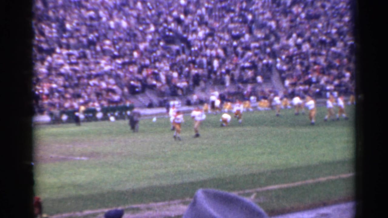 Football players on a field before packed stadium stands.