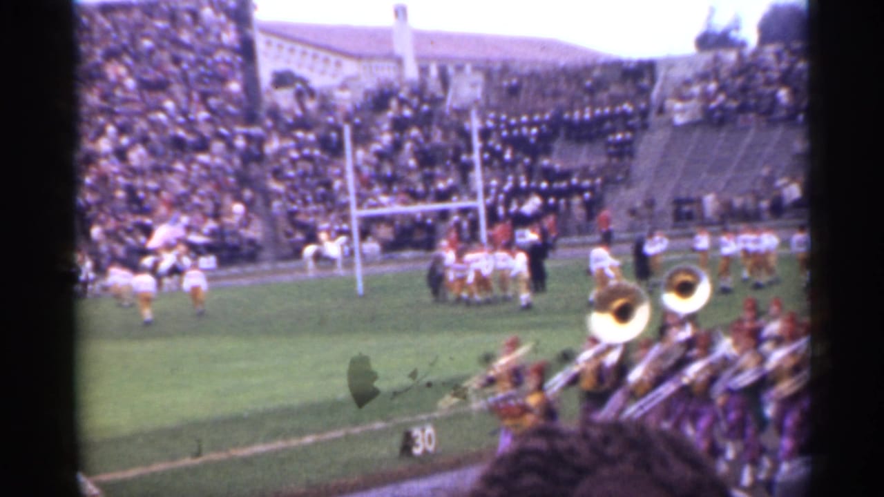 Football players near goalposts as a marching band plays in front of packed stadium stands.