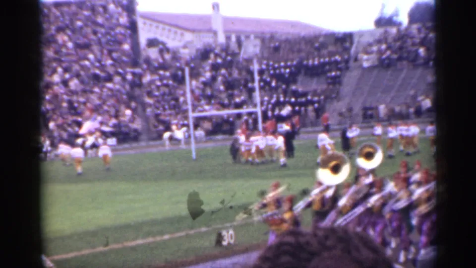 1944 San Francisco 8mm frame showing football players by the goalposts, a brass marching band, and crowded bleachers from a spectator viewpoint.