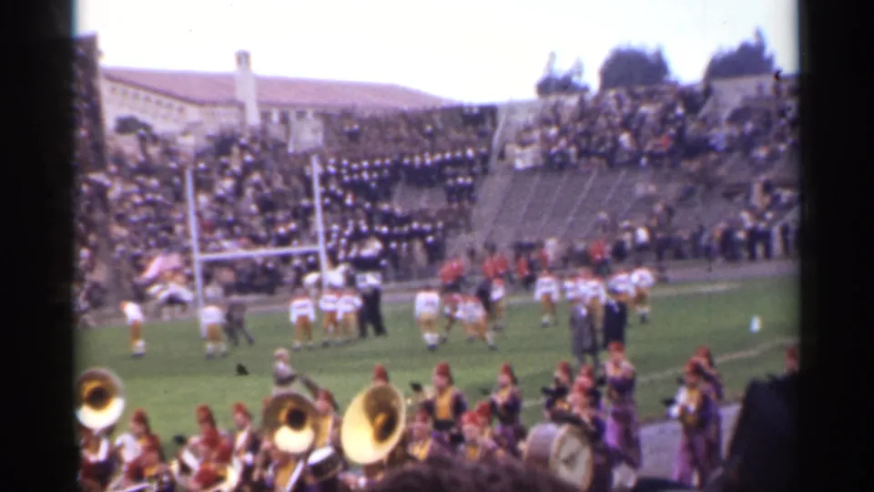 1944 San Francisco home-movie frame of a football game near the goalposts, with marching band musicians in front and crowded concrete stands beyond.