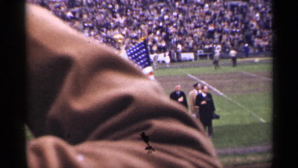 Handheld 8mm stadium view in San Francisco with an American flag, field figures, packed stands, and a foreground obstruction.